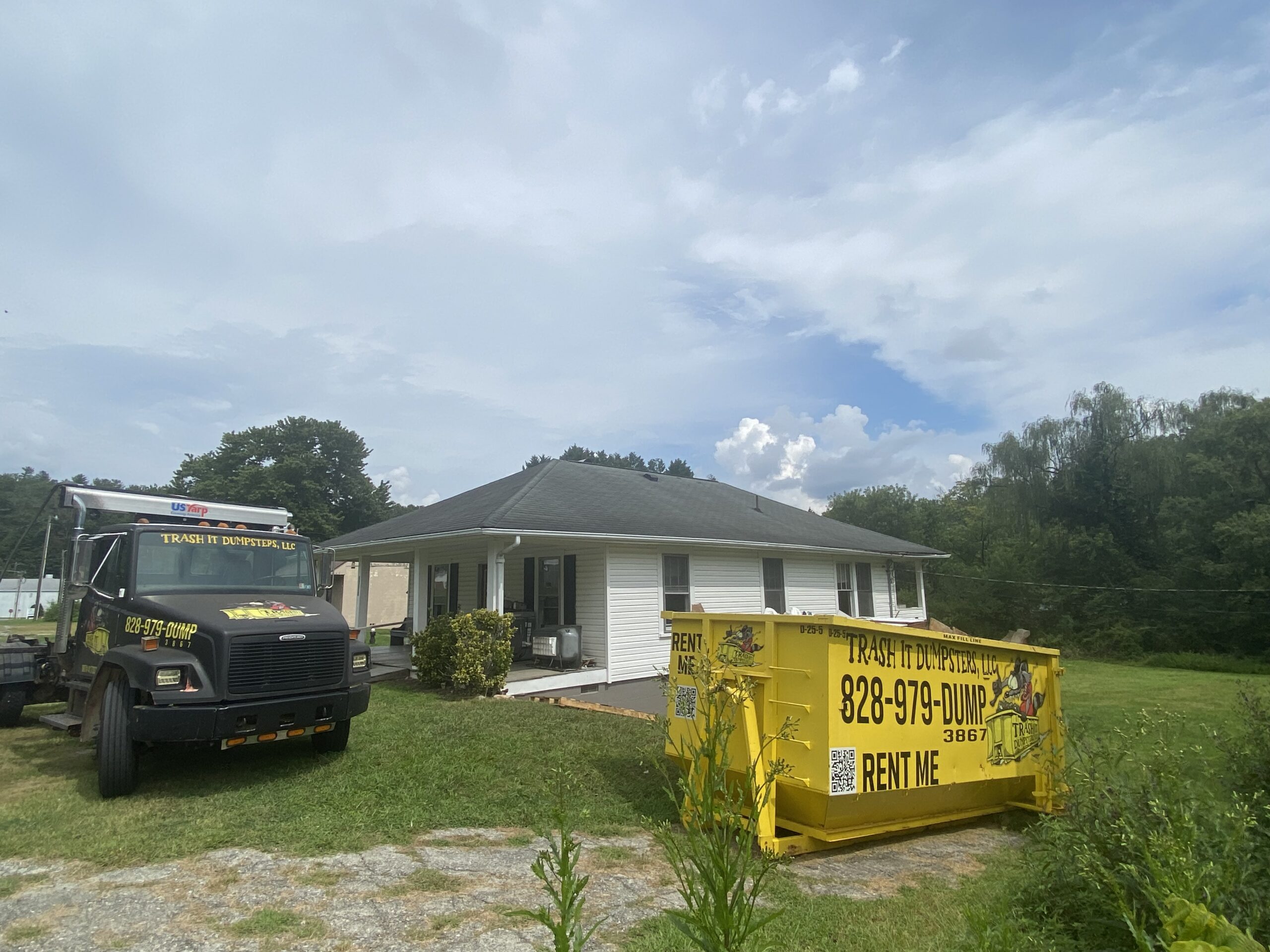 A white single-story house with a yellow 25-yard dumpster rental and a truck labeled “Trash It” parked on the grassy lawn under a partly cloudy sky.
