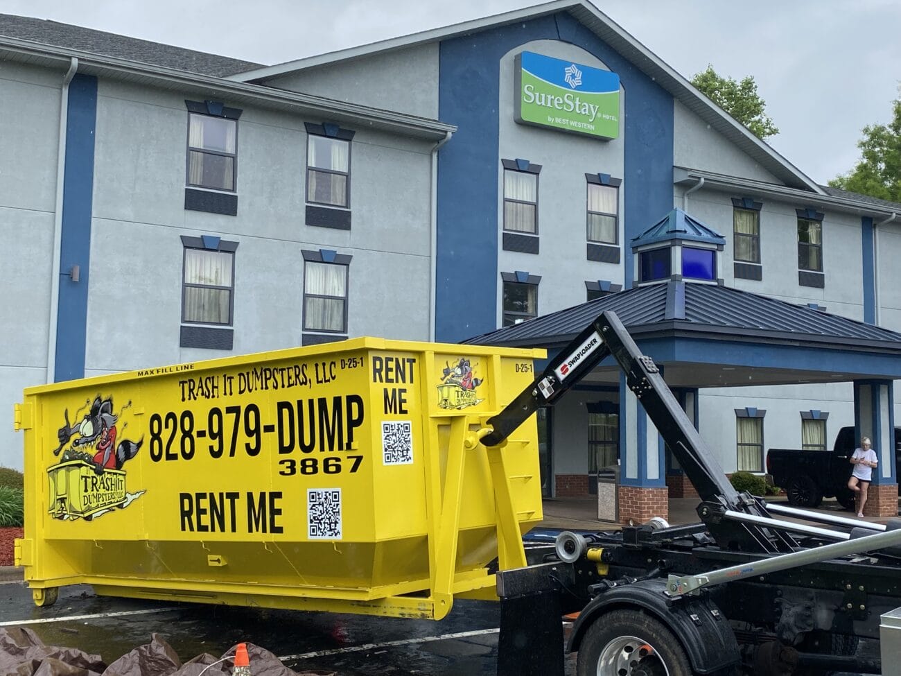 A large yellow dumpster labeled Rent Me is being delivered in front of a SureStay hotel building