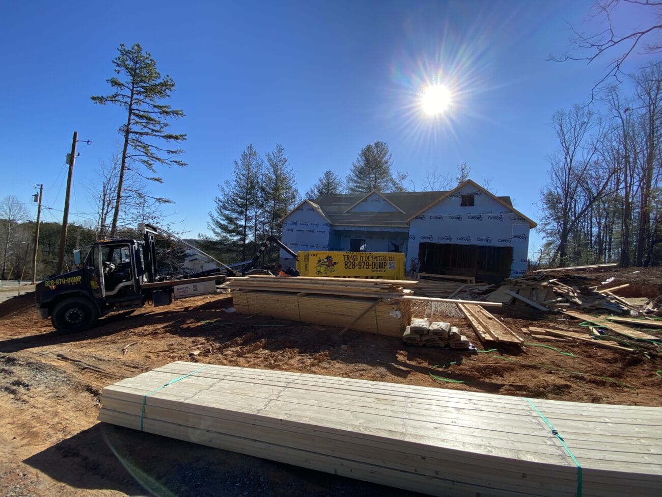 A house under construction with stacks of lumber and building materials in the yard; a forklift is unloading supplies under a bright sun