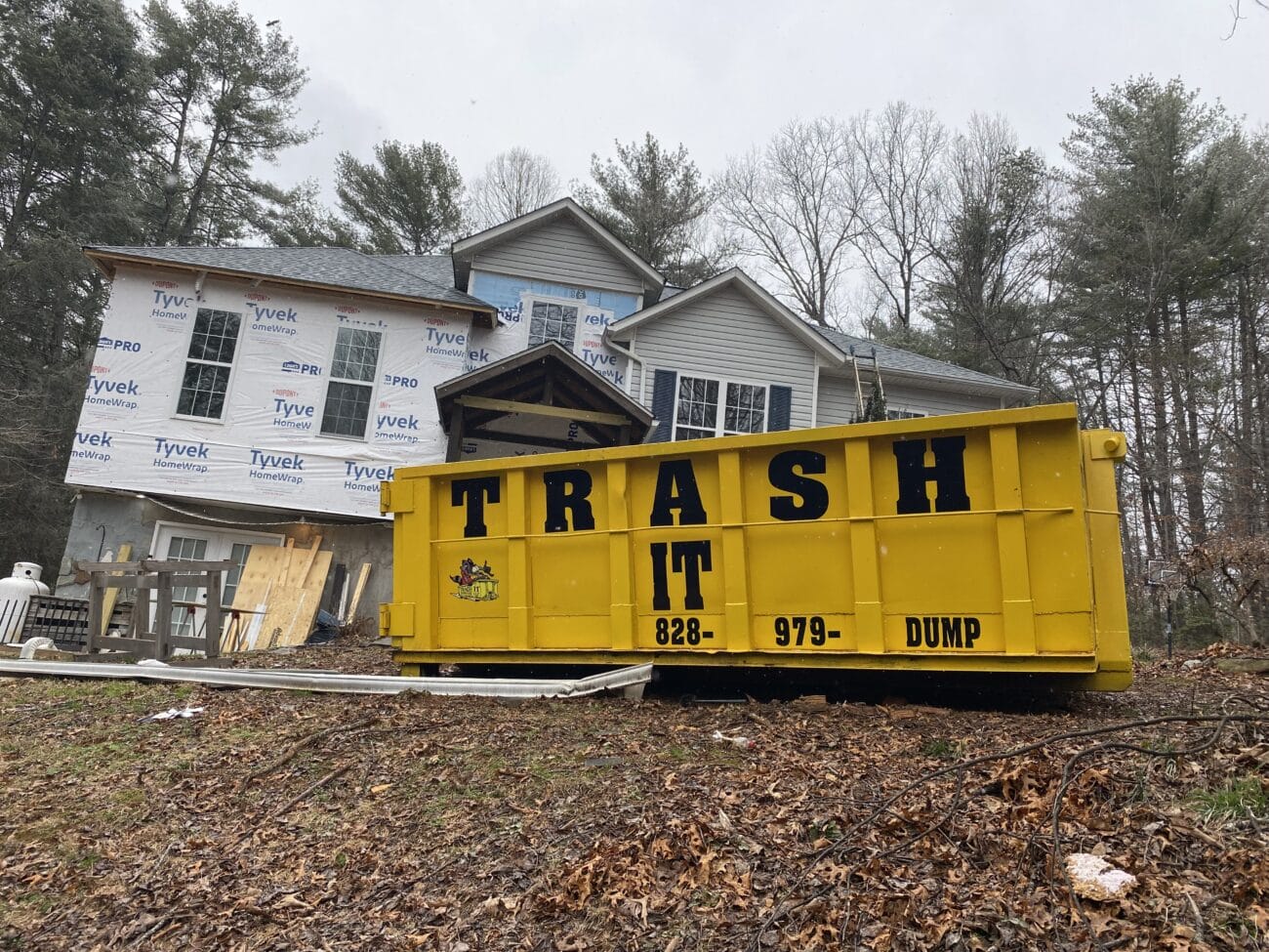 A yellow dumpster labeled TRASH IT sits in front of a partially constructed house with Tyvek wrap visible surrounded by trees and construction materials