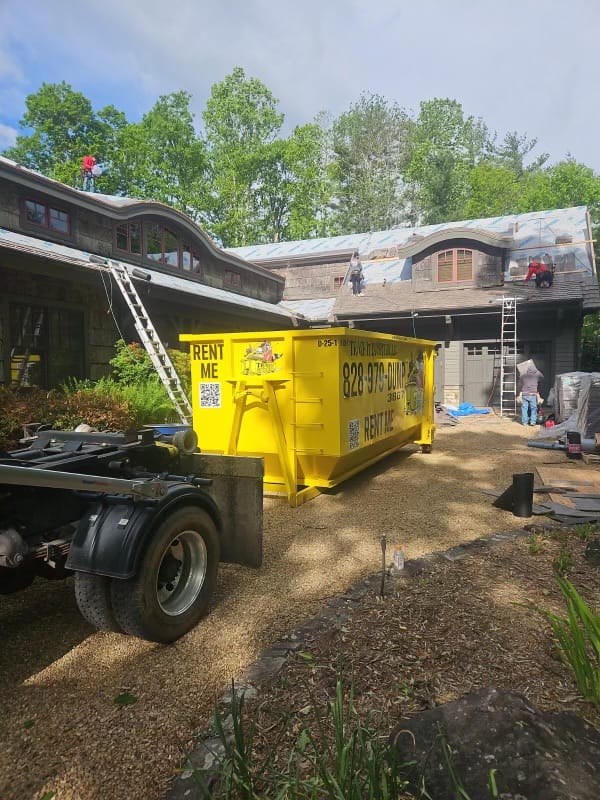 Workers installing roofing material on a house with ladders and a large yellow dumpster labeled Rent Me in the driveway A truck is parked next to the dumpster