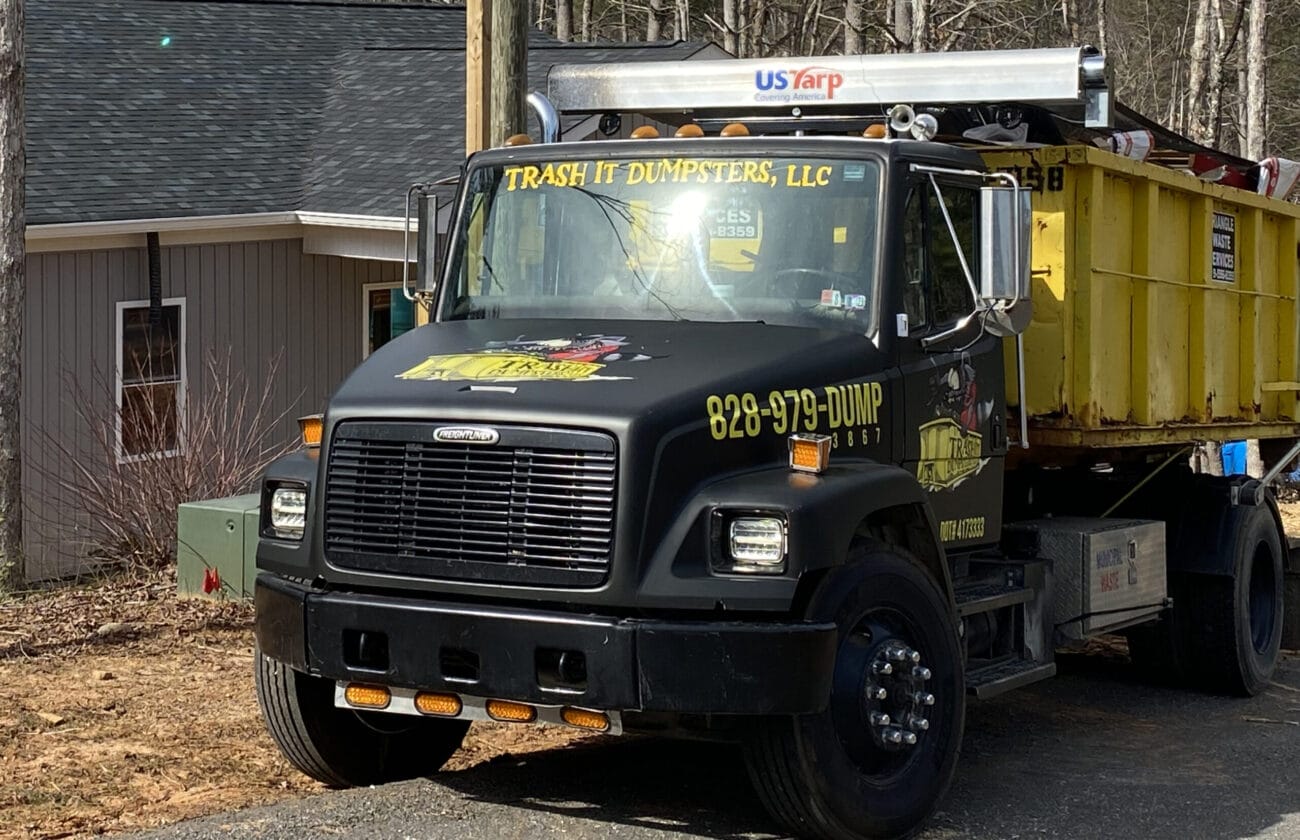 A dump truck parked on a wooded road near a house, loaded with a yellow dumpster labeled 