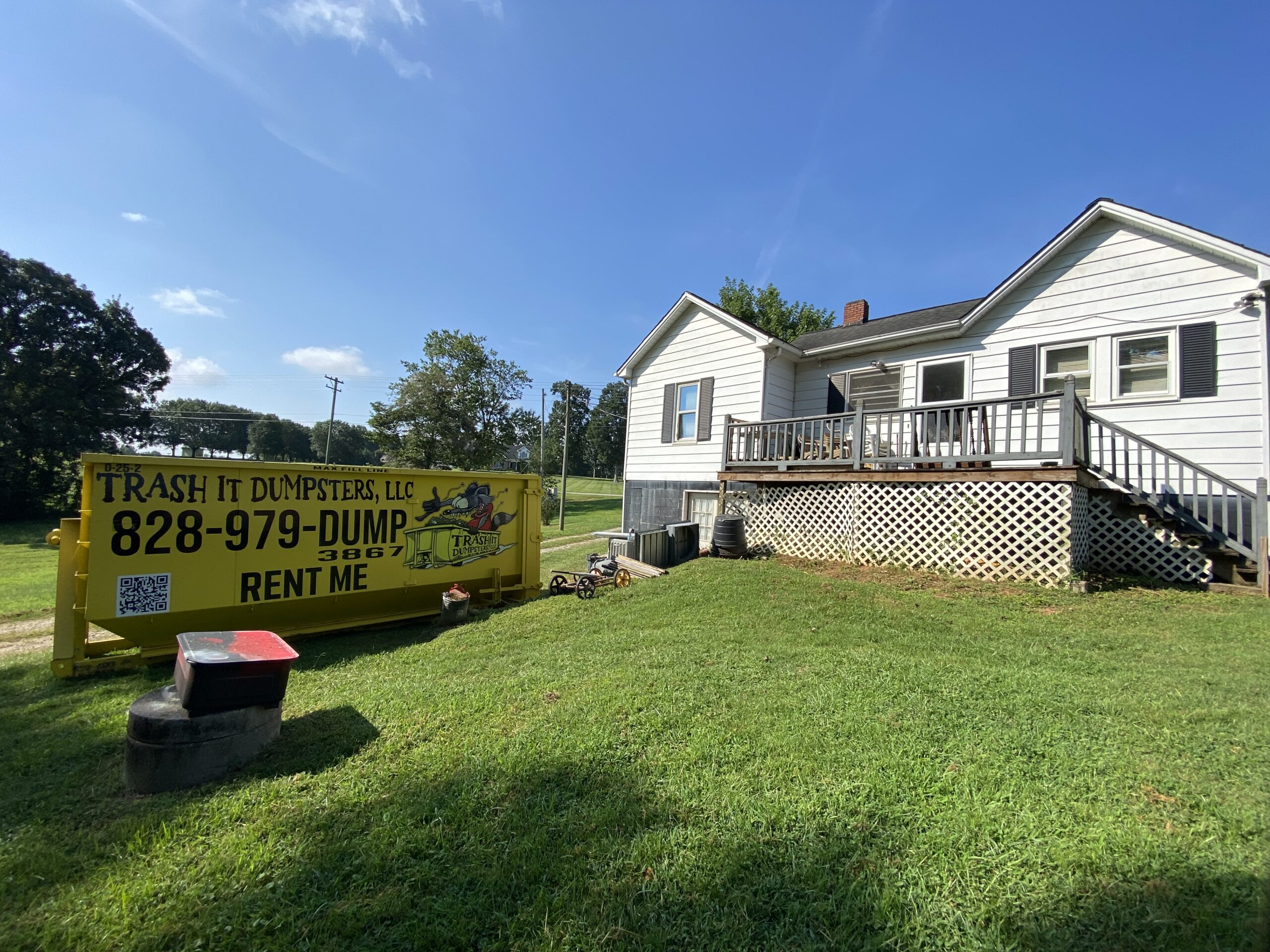 A yellow 25-yard dumpster rental labeled “Trash It Dumpsters” sits on the grass beside a white house with a deck under a clear blue sky.