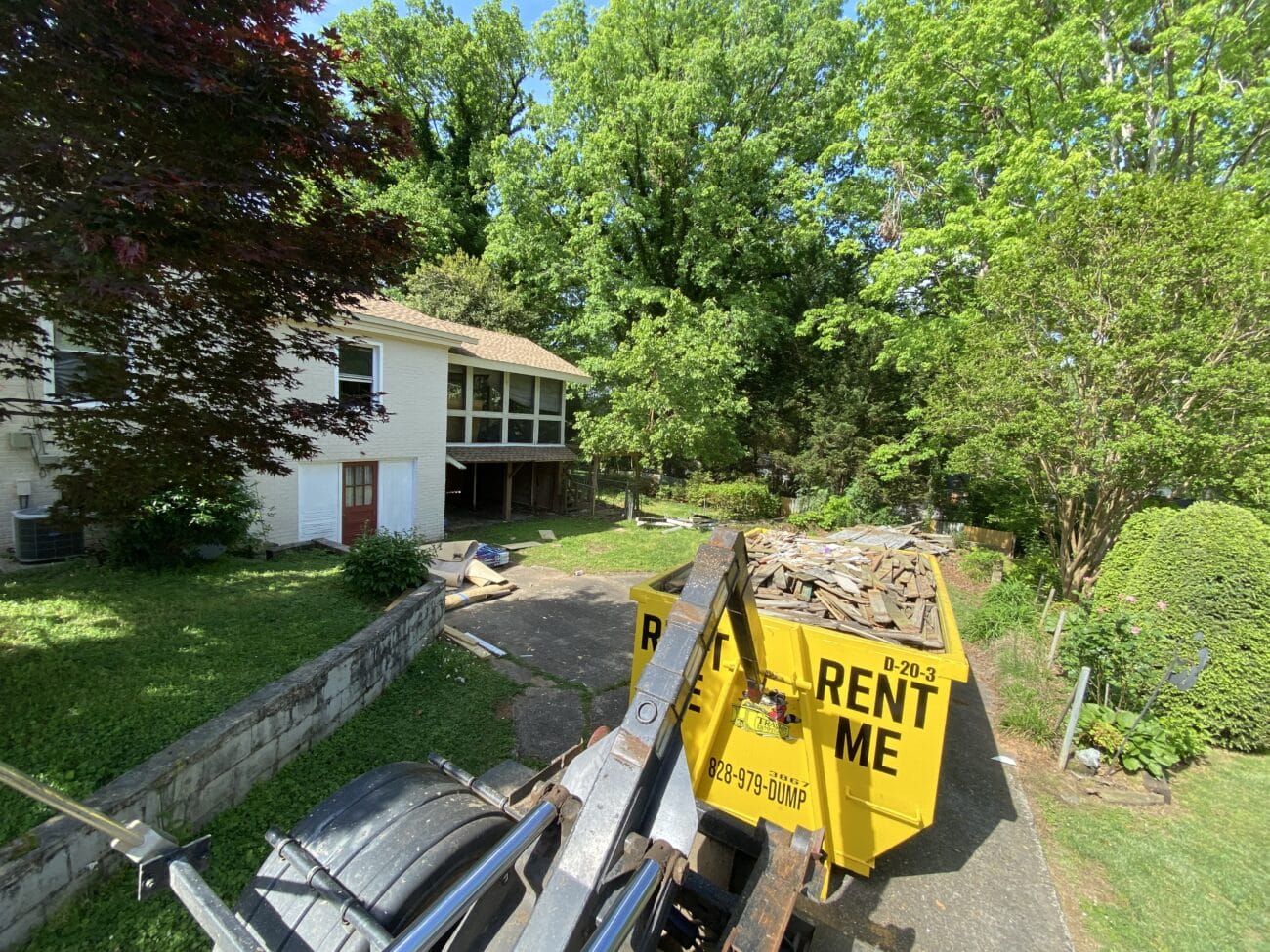 A yellow dumpster filled with debris sits in a driveway outside a white house surrounded by trees and greenery on a sunny day.