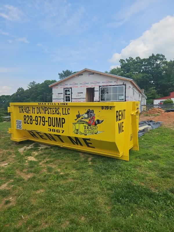 A large yellow dumpster labeled Rent Me sits in front of a partially constructed house. Construction materials are visible around the site