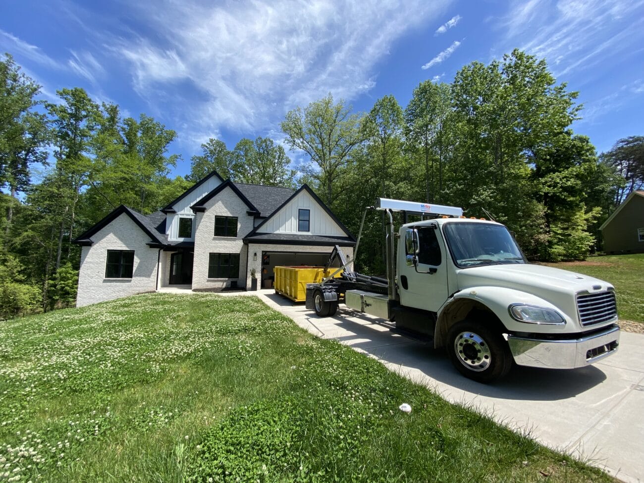 A white truck is parked on the driveway of a modern two-story house with a yellow dumpster nearby; trees and greenery surround the home.